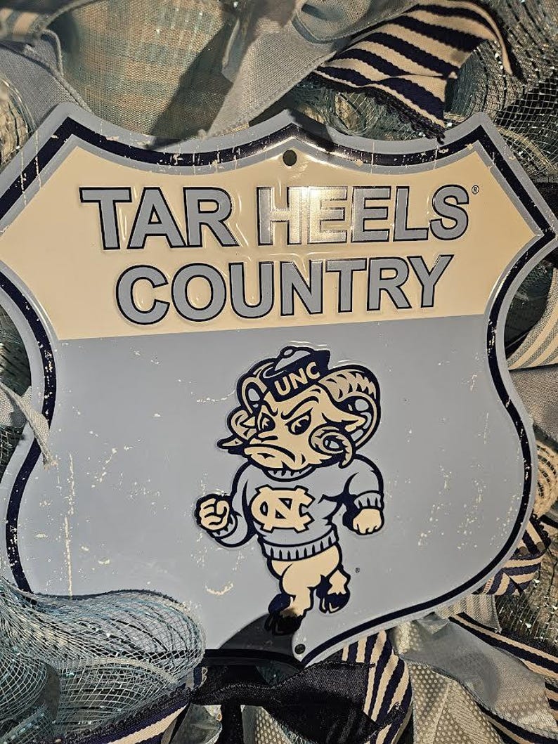Pu&ograve; includere: Un cartello in metallo azzurro con le parole "TAR HEELS COUNTRY" in grassetto. Il cartello presenta un ariete dei cartoni animati con un maglione UNC. Il cartello ha la forma di uno scudo e ha un aspetto invecchiato.