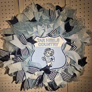 Pu&ograve; includere: Una ghirlanda decorativa con nastri e rete blu chiaro, blu navy e bianchi. La ghirlanda ha un cartello centrale con la scritta "TAR HEELS COUNTRY" e una mascotte di montone dei cartoni animati. Una decorazione festosa.