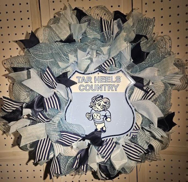 Pu&ograve; includere: Una ghirlanda decorativa con nastri e rete blu chiaro, blu navy e bianchi. La ghirlanda ha un cartello centrale con la scritta "TAR HEELS COUNTRY" e una mascotte di montone dei cartoni animati. Una decorazione festosa.