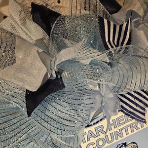 Pu&ograve; includere: Una ghirlanda decorativa con nastri in rete blu chiaro, blu navy e argento. Rifinita con fiocchi e tessuto a righe, la ghirlanda ha un aspetto strutturato. Il cartello nell'immagine recita "TAR HEEL COUNTRY".