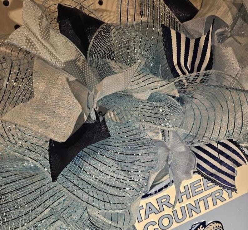 Pu&ograve; includere: Una ghirlanda decorativa con nastri in rete blu chiaro, blu navy e argento. Rifinita con fiocchi e tessuto a righe, la ghirlanda ha un aspetto strutturato. Il cartello nell'immagine recita "TAR HEEL COUNTRY".