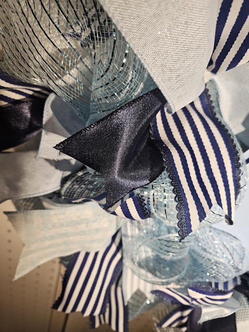 Pu&ograve; includere: Primo piano di una ghirlanda decorativa con un mix di nastri blu e bianchi, rete e tessuto. La ghirlanda ha un tema nautico con nastri a righe blu navy e bianchi e accenti blu e argento pieni. Il design &egrave; stratificato e strutturato.