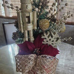 May include: A festive Christmas centerpiece featuring a gold glittered nutcracker figurine, red poinsettias, and a gold lace bow. The arrangement includes decorative sprigs and ornaments, creating a holiday display.