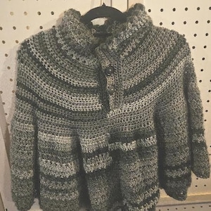 May include: A crochet poncho featuring a variegated color palette of dark green, light green, gray, and black. The garment has a high collar with a buttoned placket containing two dark buttons, and a textured, ribbed stitch pattern throughout.
