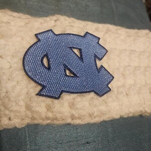 May include: Close-up of a blue and white crocheted item. A blue emblem with the letters "NC" is centered on a white crocheted band. The background is a light blue fabric. The item appears to be a headband or similar accessory.