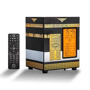 May include: A black and gold decorative clock shaped like the Kaaba, with a digital display showing the time, date, and temperature. A remote control is on the left. The clock has gold accents and Arabic script.