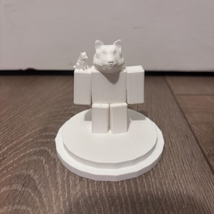 May include: A white 3D-printed figurine of a block-shaped figure with a cat's head and a smaller cat figure on its shoulder. The figurine is mounted on a circular base with tiered edges. The overall design is minimalist and modern.