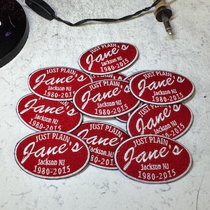 May include: A group of red and white oval patches. Each patch displays the text "JUST PLAIN Jane's Jackson NJ 1980-2015" in a white, cursive font. The patches are laid out on a light-coloured surface.