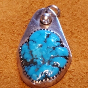 May include: A turquoise pendant with a silver setting. The turquoise is a bright blue with dark veins running through it. The pendant is oval shaped and has a silver bail at the top.