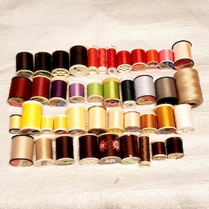 May include: A collection of assorted sewing thread spools in various sizes and colors, including shades of black, white, red, yellow, brown, green, blue, and pink. The spools are arranged in four horizontal rows, featuring different brands.