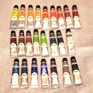May include: A collection of gouache paint tubes in various colors, including yellow, orange, red, green, blue, and brown. Each tube has a black cap and is labeled with the brand name. The tubes are arranged on a light-colored surface.