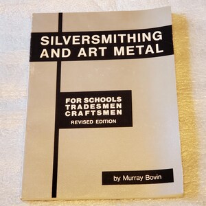 May include: A softcover book titled Silversmithing and Art Metal for Schools Tradesmen Craftsmen Revised Edition by Murray Bovin. The cover is light gray with black horizontal and vertical bars, featuring white text throughout the front cover design.