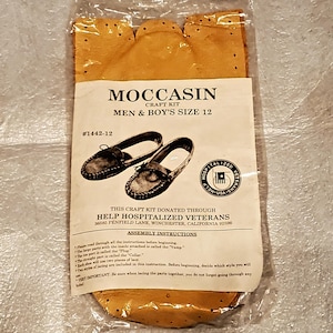 May include: A tan leather moccasin craft kit in a clear plastic bag. The label reads MOCCASIN CRAFT KIT MEN & BOY'S SIZE 12 #1442-12, featuring an illustration of two moccasins and text about Help Hospitalized Veterans and assembly instructions.