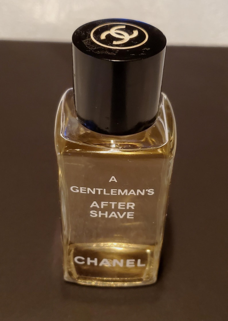 Może przedstawiać: Mała, prostokątna szklana butelka wody po goleniu Chanel. Butelka ma czarną nakrętkę ze złotym logo. Z przodu widnieje napis "A Gentleman's After Shave".