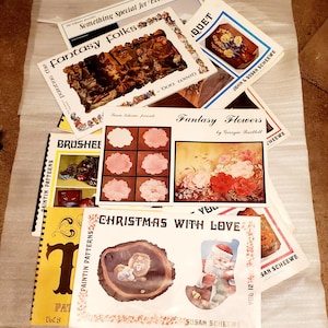 May include: A collection of vintage art books and patterns, featuring titles like "Fantasy Folks" and "Christmas with Love." The covers display illustrations of fantasy scenes, flowers, and holiday themes. The books are in various sizes and colors.