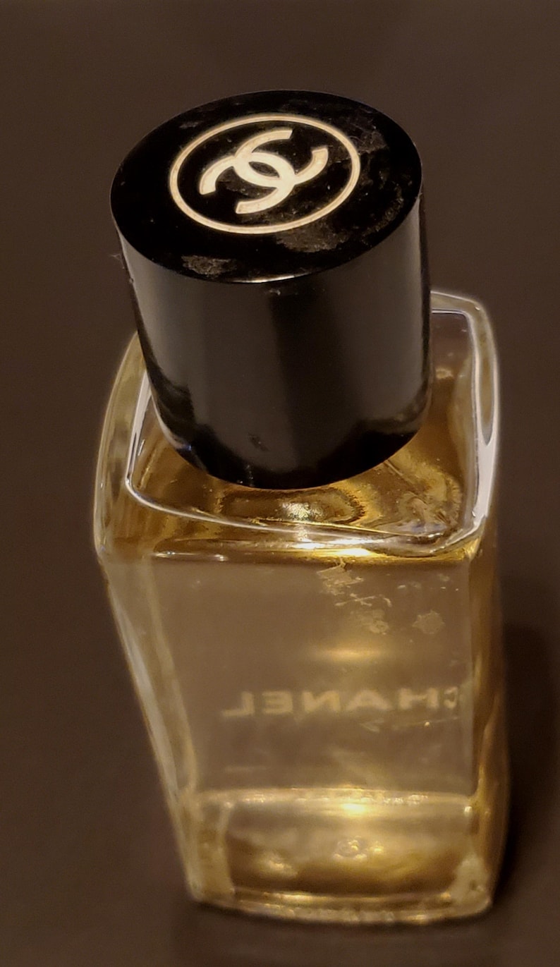 Może przedstawiać: Mały, przezroczysty flakon perfum z czarną nakrętką. Nakrętka posiada białe logo. Butelka zawiera złoty płyn. Słowo "CHANEL" jest wydrukowane odwrotnie na przodzie butelki.
