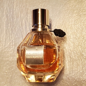 May include: A clear, faceted perfume bottle with a gold cap and a black flower-shaped charm. The bottle contains a golden-colored liquid and features a silver label with the text "FLOWERBOMB VIKTOR&ROLF".