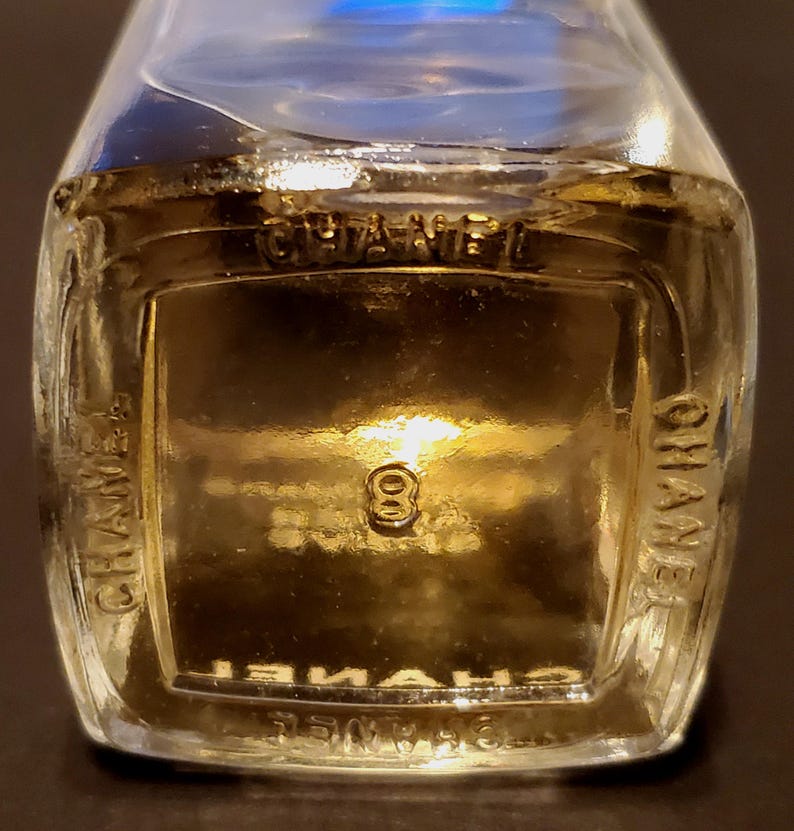 Może przedstawiać: Zbliżenie na przezroczysty szklany flakon perfum z wytłoczonym napisem "CHANEL" po bokach. Numer "8" jest widoczny na środku butelki. Płyn wewnątrz ma złoty odcień.