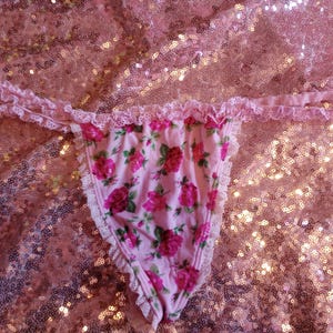 May include: Pink thong underwear with a floral rose pattern. The underwear has a delicate lace trim along the edges and straps. The background is a shimmering pink sequined fabric.