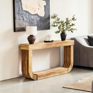May include: A light-colored wooden console table with a curved base and a rectangular top. A small lamp with a white shade and a dark brown base sits on the table, along with a black vase and a small tray with candles. The table is in a living room setting.