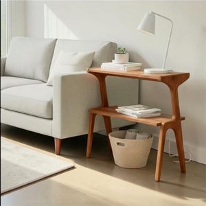 May include: A two-tiered wooden side table with a unique, curved design. The table has a natural wood finish and features a lower shelf and a woven basket. A white lamp and a small potted plant sit on top. A light gray sofa is in the background.