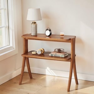 May include: A two-tiered wooden side table with a warm brown finish. A white lamp with a patterned base, a black alarm clock, and other decorative items sit on the top shelf. The lower shelf holds headphones and books.