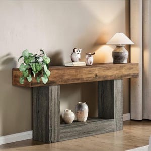 Op de afbeelding: Een rustieke houten console met een donkerbruin blad en een grijze basis. De tafel is versierd met een kleine potplant, uilbeeldjes, een lamp en twee decoratieve vazen. De tafel is van hout.