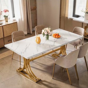Op de afbeelding: Een rechthoekige eettafel met een wit marmerlook blad en goudkleurige gebogen poten. Zes beige gestoffeerde stoelen omringen de tafel. Een vaas met bloemen, een schaal met sinaasappels en een kan sap staan op de tafel.