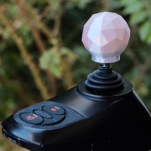 May include: A black joystick control panel with a geometric, pink and white sphere-shaped knob. The control panel has several buttons with red and blue accents. The background is blurred, showing green foliage.