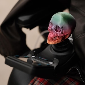 May include: A colorful skull-shaped gear shift knob. The skull is painted with a gradient of colors, including green, pink, and orange. The knob is mounted on a black base, and the background includes a black jacket and plaid fabric.