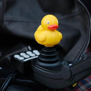Könnte beinhalten: Eine gelbe Quietscheente mit rotem Schnabel und schwarzen Augen sitzt auf einem schwarzen Joystick. Die Ente ist der Mittelpunkt, vor einem verschwommenen Hintergrund. Der Joystick ist Teil eines Steuerungssystems.