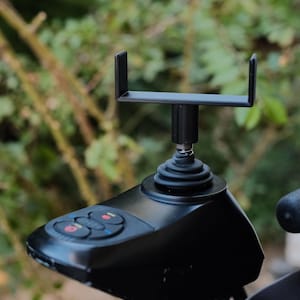 Puede incluir: Primer plano de un panel de control de joystick negro con botones rojos y azules. El panel de control tiene un diseño curvo y está montado en un poste negro. Un soporte negro está unido a la parte superior del poste.