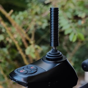 May include: A black joystick controller with a ribbed handle and a base with red buttons. The controller is mounted on a black support, with a blurred green background of foliage.