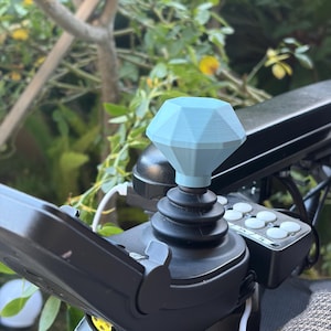 Könnte beinhalten: Ein hellblaues, rautenförmiges Joystick-Knopf ist an einem schwarzen Bedienfeld befestigt. Der Knopf besteht aus einem 3D-gedruckten Material. Der Hintergrund zeigt grünes Laub und einen Blick auf eine sonnige Umgebung im Freien.
