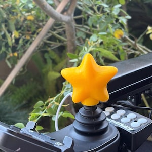 May include: A vibrant yellow star-shaped knob is mounted on a black joystick control. The joystick is part of a mobility aid, with a panel of white buttons and other controls visible. The background is blurred with green leaves.