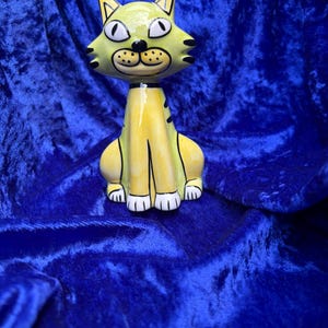 May include: A whimsical, hand-painted ceramic cat figurine. The cat has a yellow body with black stripes and white paws. The head is a lighter shade of yellow with black and white facial features. The cat is sitting upright.