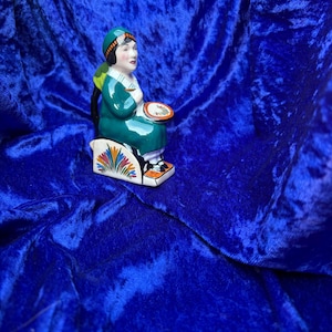 Peut inclure: Figurine en céramique d'une femme assise, vêtue d'une robe et d'un chapeau verts, tenant une assiette. La figurine a une base blanche avec des motifs floraux colorés. Le fond est un tissu bleu foncé.