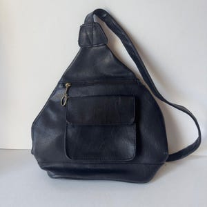 May include: A black triangular sling backpack featuring a single adjustable shoulder strap, a horizontal zipped compartment with a metallic ring pull, and a central front pocket with a flap closure. The bag has a smooth, textured surface.