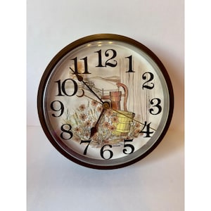 May include: A round wall clock with a dark brown frame and a clear glass cover. The clock face features black Arabic numerals and a central illustration of a water pump, a wooden bucket, and orange flowers in a field under a pale sky.