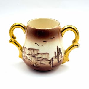 May include: A ceramic sugar bowl with two gold-toned handles and a cream-colored body. The exterior features a brown gradient finish with a painted desert landscape, including a saguaro cactus, rocky formations, and birds in flight.