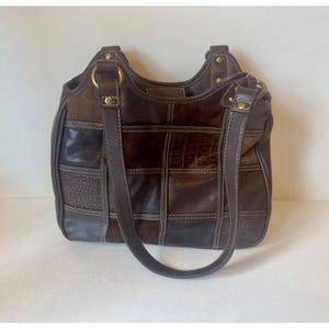 Puede incluir: Un bolso de mano marrón oscuro con un diseño de patchwork y texturas variadas, incluyendo paneles de cuero liso, granulado y con relieve de cocodrilo. El bolso tiene dos correas de hombro sujetas con anillos y remaches de metal latonado.