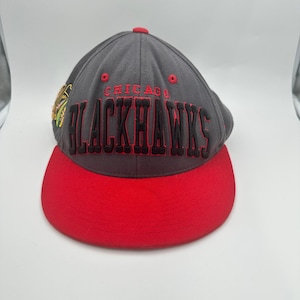 Könnte beinhalten: Eine graue Baseballkappe mit rotem Schirm, die auf der Vorderseite den in Schwarz mit roten Umrandungen gestickten Schriftzug CHICAGO BLACKHAWKS zeigt. Ein kleines, rundes Team-Logo-Emblem ist an der Seite der Kappe mit roten Ösen zu sehen.