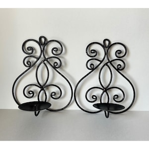 May include: A pair of black metal wall sconces featuring intricate scrollwork designs. Each sconce has a circular candle plate at the base supported by a curved frame with symmetrical loops and swirls extending upward in a decorative pattern.