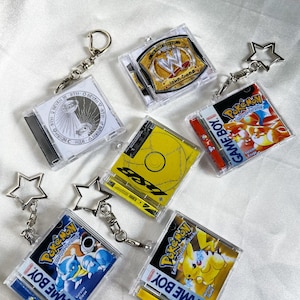 May include: A selection of clear plastic keyrings with diverse designs. Some keyrings feature Pokémon game covers, while others display a wrestling championship belt and abstract art. Each keyring has a silver star or round clasp.