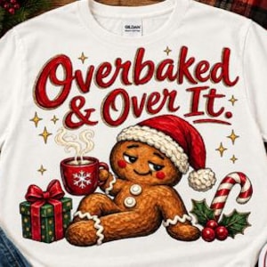 May include: A white t-shirt featuring a graphic of a gingerbread person wearing a Santa hat, holding a red mug with a snowflake design, next to a green gift box with a red bow and a candy cane, with the text Overbaked & Over It in red.