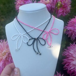 May include: Three beaded bow necklaces in white, black, and pink displayed on a mannequin. The necklaces have delicate bow designs with small beads and dangling accents. The background features pink flowers and greenery.