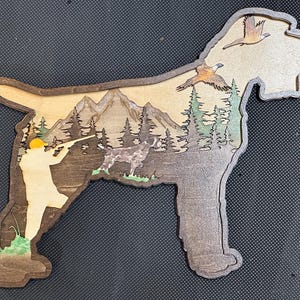 May include: Wooden dog-shaped wall art depicting a hunting scene. The artwork shows a hunter, mountains, trees, and birds in flight. The piece is made of layered wood with natural and painted colours.