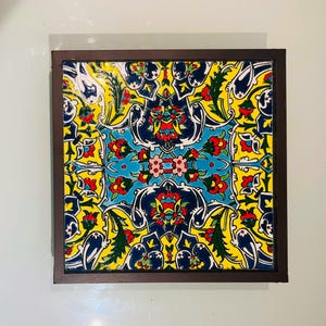 May include: A square ceramic tile framed in dark brown wood, featuring a symmetrical floral pattern with vibrant yellow, deep blue, turquoise, red, and green colors. The intricate design displays stylized blossoms and leaves in a repeating layout.
