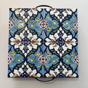 May include: Square serving tray with a floral tile design. The tiles feature a blue, white, green, yellow, and red pattern. The tray has a black metal handle on each side.