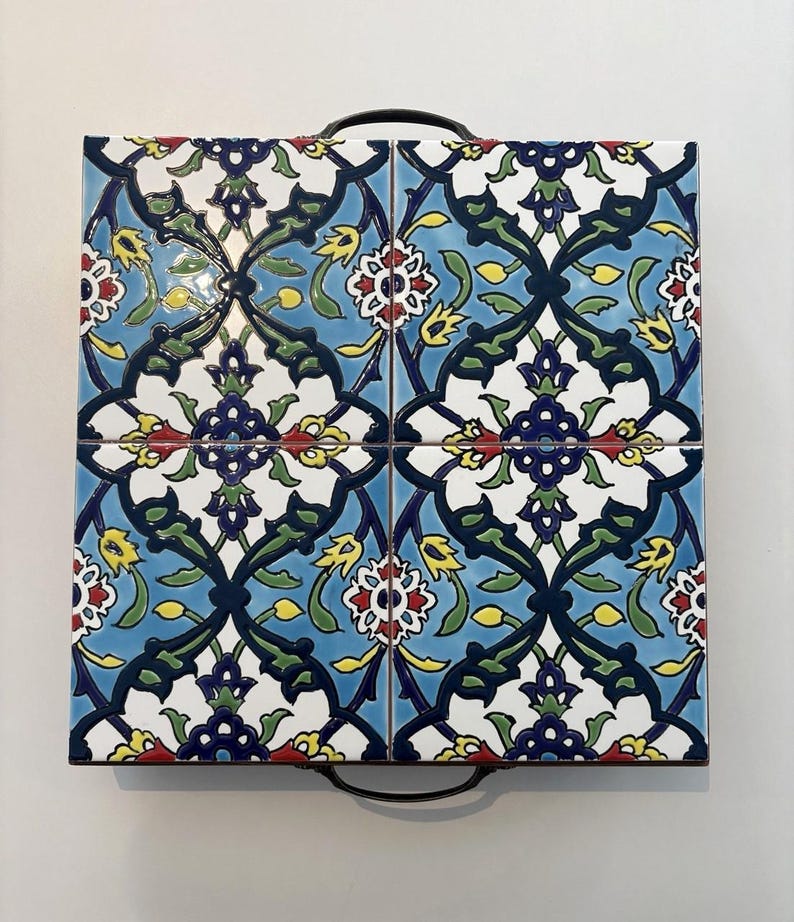 May include: Square serving tray with a floral tile design. The tiles feature a blue, white, green, yellow, and red pattern. The tray has a black metal handle on each side.